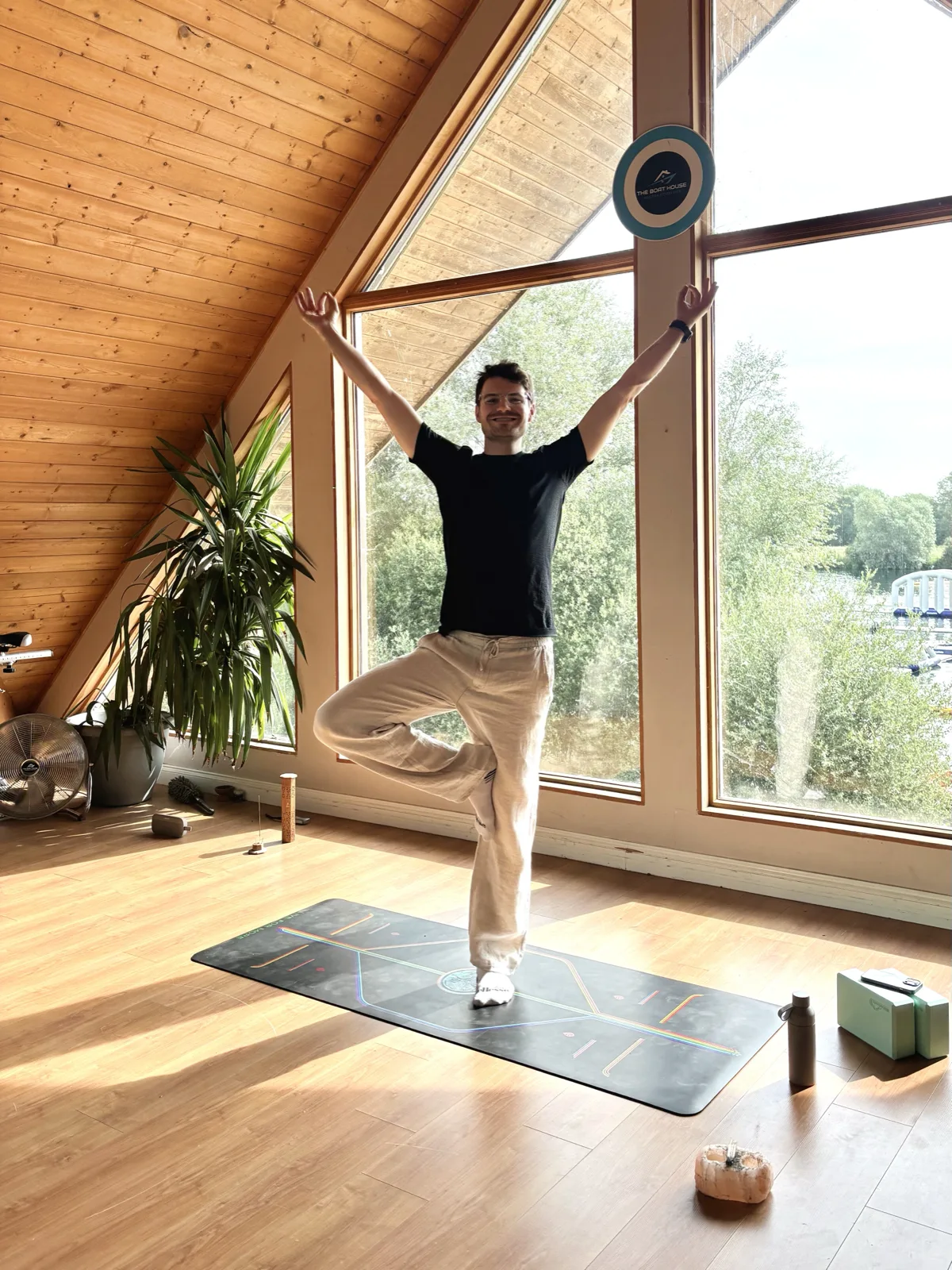 Kai balancing in tree pose on a mat, arms raised overhead, in a bright loft studio with a pitched wooden ceiling and tall windows looking out over trees and water.
